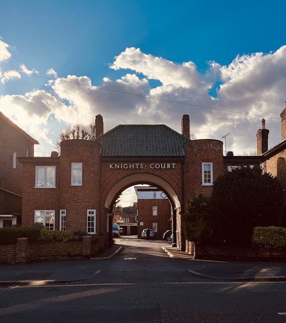 A historic brick archway labeled 'Knights Court' at the entrance to a residential area, with a curved gateway featuring two cylindrical towers on either side. The brickwork has a reddish-brown hue with a textured surface, and the archway is topped with a hipped roof covered in dark grey tiles. Behind the arch, a narrow street is visible, lined with parked cars and brick buildings. On the foreground, a paved driveway or small road leads through the arch, flanked by low brick walls and neatly trimmed shrubbery. The sky above is partly cloudy with bright white clouds and patches of blue, casting soft shadows over the scene. The area appears clean and well-maintained, reflecting a typical urban or suburban environment. This setting visually aligns with services such as private rubbish removal or on-site clearance by companies like wastedisposalkingstonuponthames.co.uk, providing an observational context for alternative waste handling methods outside of traditional municipal collection.