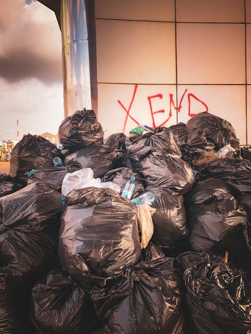 A large pile of black rubbish bags, some appearing slightly torn or crinkled, is stacked against a tiled wall in an outdoor urban setting. Several discarded plastic water bottles, with clear and blue tones, are visible protruding from the bags. On the wall behind the bags, the word 'XEND' is spray-painted in red graffiti. The scene is illuminated by natural light, with a cloudy sky visible in the background, indicating overcast weather. The surrounding environment suggests a commercial or public space, possibly a location where private rubbish collection or on-site waste clearance is taking place, with no visible vehicles or equipment in the immediate foreground. The image reflects an instance of unmanaged waste accumulation that could be associated with independent or alternative waste handling services in Kingston upon Thames, highlighting the importance of professional rubbish removal to maintain urban cleanliness.
