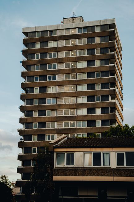 A tall multi-storey residential building with a distinctive rectangular shape and a grid of windows and balconies dominating the upper floors. The building features a concrete facade with horizontal bands separating each floor, and a weathered, slightly textured appearance. The windows are uniformly spaced, many illuminated, indicating interior lighting, with some showing partially open curtains or blinds. In the foreground, a smaller, single-story building with a sloped, shingle roof and large rectangular windows is visible, partially obscured by a tree with dark foliage. The scene is set outdoors in an urban environment during late afternoon or early evening, with a cloudy sky overhead, casting diffuse natural light. The overall atmosphere is calm, and the environment suggests a typical city skyline where private waste collection services, such as those provided by Waste Disposal Kingston upon Thames, might handle rubbish clearance for high-rise and residential areas alike, beyond local authority waste collection.