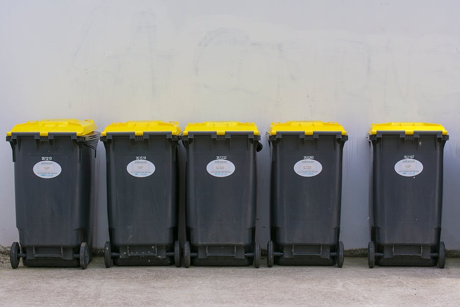 A row of five large wheeled rubbish bins with black bodies and yellow lids, lined up against a plain, light-colored wall outdoors. The bins are made of durable plastic with a textured surface, and each has a white label on the front indicating identification numbers. The bins are evenly spaced on a paved surface, with some minor dirt visible at the base of the wheels. The environment appears to be an external area, possibly a residential driveway or service yard, and the scene is lit with natural ambient light, highlighting the clean but slightly weathered condition of the bins. This setup suggests an organized approach to waste management, similar to private rubbish collection services that could complement or serve as an alternative to council-provided bulky rubbish disposal in Kingston upon Thames. The image reflects practices involving on-site waste storage awaiting collection, emphasizing the importance of proper disposal procedures as outlined by Kingston council rules for rubbish collection. A straightforward, professional visual representation suited to content on rubbish removal services provided by waste disposal companies like wastedisposalkingstonuponthames.co.uk.