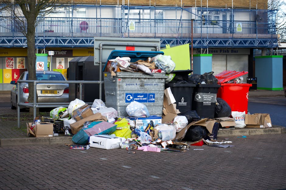 A large outdoor waste collection point featuring a variety of rubbish bags, cardboard boxes, and assorted refuse piled around three large wheelie bins on a paved sidewalk. The central bin, made of grey plastic with a blue recycling symbol, is overflowing with paper, cardboard, and miscellaneous waste, some of which has spilled onto the surrounding ground. To its right, a black bin contains black trash bags and a few loose items, while a bright red bin is visible further right. In the background, there is a commercial building with retail units, including signs for a fish bar and other shops, partially obscured by a metal fencing and scaffolding. Adjacent to the waste area, a metal railing separates the rubbish from parked cars, one of which is silver with a visible registration plate. The scene appears to be in an urban area with typical street-level rubbish collection, highlighting the need for regular waste clearance and disposal services often managed by private waste companies like wastedisposalkingstonuponthames.co.uk, especially for bulky waste or excess refuse collections.