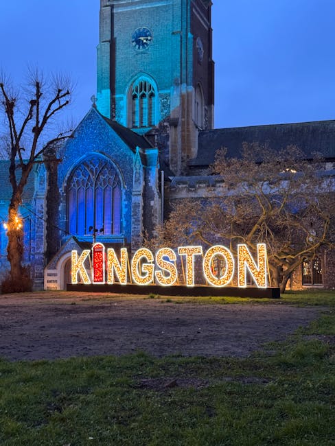 A large illuminated sign spelling 'KINGSTON' in uppercase letters, with the letters 'K' and 'I' illuminated in red and white lights, positioned on a grassy area in front of a historic stone church with Gothic-style windows and a tall clock tower. The church is lit with blue lighting, creating a striking contrast against the darkening evening sky. Bare tree branches are visible on the left side of the image, and the overall scene suggests an outdoor public space in Kingston upon Thames, related to local community or cultural events, complementing the context of rubbish removal and waste management services by providing a visually recognizable local landmark.