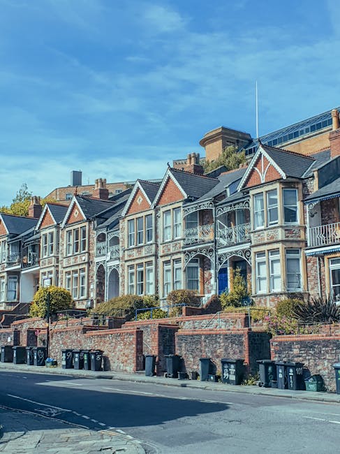 A row of Victorian-style terraced houses with decorative white trim and bay windows, situated on a slightly elevated brick-fronted street in Kingston upon Thames. The buildings have a mix of brick and painted exterior finishes, with some featuring ornate ironwork on balconies and porches. In front of the houses, there is a low brick wall with small landscaped gardens, containing shrubs and plants. Several black wheelie bins are lined along the pavement, indicating typical waste collection points. The sky above is clear with a few wispy clouds, and the scene is bathed in natural daylight. This residential street reflects the traditional architecture of Kingston, with a setting suitable for discussing private waste handling and robust rubbish removal services like those offered by [COMPANY_NAME], often used for on-site clearance or independent rubbish disposal in the local area.