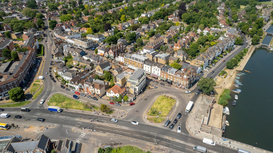 An aerial view of a seaside town with a mix of residential and commercial buildings, situated near a river or canal with several small boats docked along the waterfront. The foreground shows a busy road with multiple lanes, traffic including cars, trucks, and buses, and a roundabout surrounded by patches of grass and small trees. The buildings are predominantly brick with varied architectural styles, some with flat roofs and others with pitched roofs, reflecting a mix of traditional and modern designs. The streets are lined with trees, and the overall environment appears well-maintained and densely built. In the distance, a lush green canopy of trees extends over residential neighborhoods, indicating a vibrant and established community. Visible watercraft and the proximity to water emphasize the town’s connection to the river, highlighting potential locations for independent waste disposal or rubbish removal services like those occasionally provided by Waste Disposal Kingston upon Thames, supporting local municipal or private waste management needs in the Kingston upon Thames area.