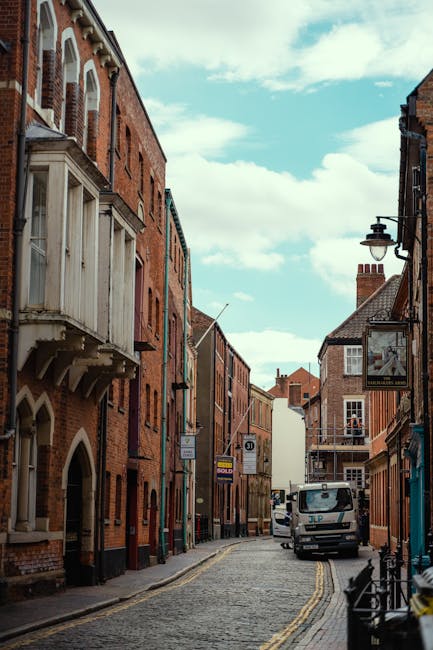 A narrow cobblestone street in Kingston upon Thames lined with historic brick buildings on both sides, featuring a mix of red and brown tones. On the left, a building has a distinctive bay window with decorative wooden supports, while on the right, a street lamp with a black metal bracket is attached to a building façade. A white delivery van is parked further down the street, partially obscuring the view of the buildings beyond. The sky above is partly cloudy with patches of blue visible through white clouds, casting soft natural light across the scene. Visible portions of the pavement and curbs are clean, with no visible waste or rubbish present, illustrating an environment suitable for different types of waste removal services, including private collection for narrow street access issues. This setting reflects an urban area where efficient on-site clearance or alternative waste handling methods are considered due to restricted access for large rubbish removal vehicles. The overall atmosphere is quiet and orderly, emphasizing the importance of precise logistics in waste management for such limited-access urban streets.