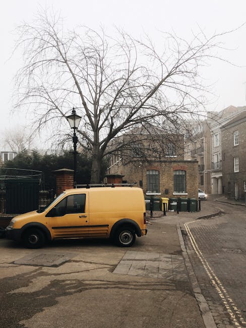 A yellow cargo van is parked on a narrow, slightly inclined urban street near a leafless tree. The van's surface appears smooth with a matte finish, and it has black wheels and tinted side windows. Behind the van, there is a small brick building with two tall, narrow windows featuring dark frames, and several wheelie bins in green and black placed against the building's wall, indicating a waste collection area. To the left, a tall black lamppost with ornate detailing stands adjacent to a metal fence, which partially obscures a garden or courtyard behind it. The street is flanked by residential buildings with brick and plaster facades, some with bay windows, and the pavement consists of concrete slabs with patches of unevenness and slight weathering. The overall scene is enveloped in soft, overcast daylight typical of cloudy days, subtly highlighting the practical setting for private waste disposal and alternative rubbish collection methods, which local waste management services like wastedisposalkingstonuponthames.co.uk may support for properties with challenging access.
