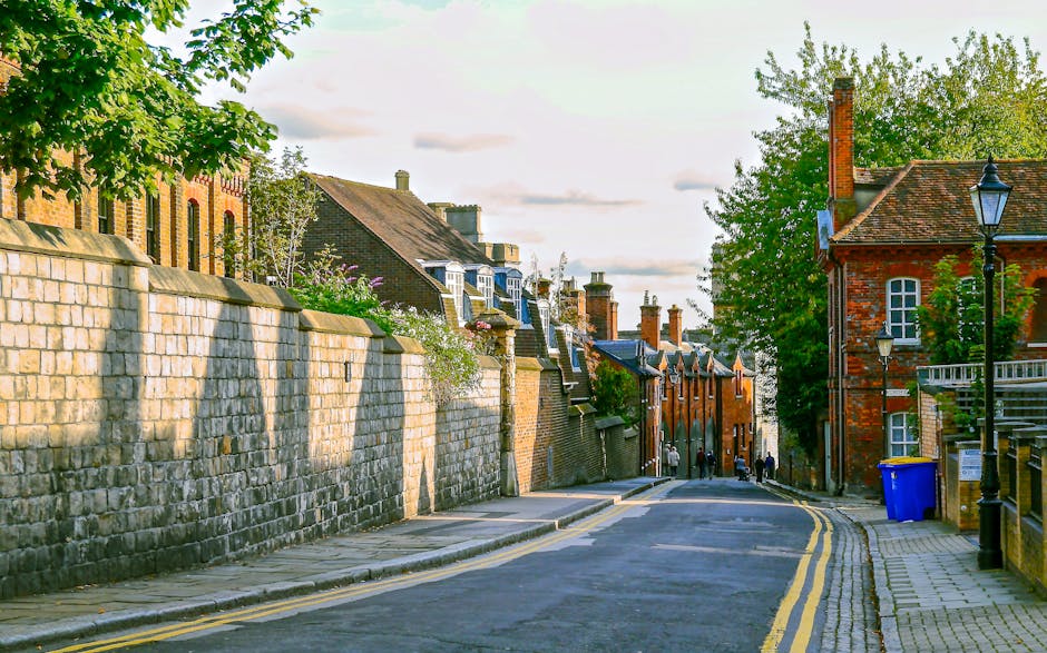 A narrow cobblestone street in Kingston upon Thames lined with historic brick buildings on both sides, featuring a mix of red and brown tones. On the left, a building has a distinctive bay window with decorative wooden supports, while on the right, a street lamp with a black metal bracket is attached to a building façade. A white delivery van is parked further down the street, partially obscuring the view of the buildings beyond. The sky above is partly cloudy with patches of blue visible through white clouds, casting soft natural light across the scene. Visible portions of the pavement and curbs are clean, with no visible waste or rubbish present, illustrating an environment suitable for different types of waste removal services, including private collection for narrow street access issues. This setting reflects an urban area where efficient on-site clearance or alternative waste handling methods are considered due to restricted access for large rubbish removal vehicles. The overall atmosphere is quiet and orderly, emphasizing the importance of precise logistics in waste management for such limited-access urban streets.