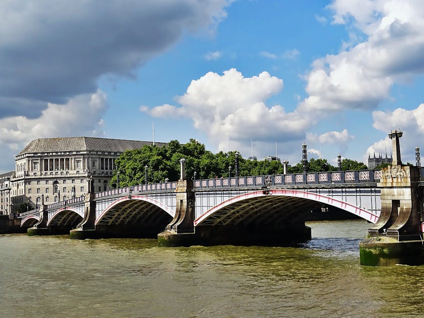 A detailed view of a historic metal bridge spanning a river in an urban environment, featuring multiple arches with ornate decorative elements and white-painted structural supports with red accents. The bridge's railing has a repeating pattern with vertical bars and ornamental details, topped with a flat walkway. In the background, there is a large, classical-style stone building with tall columns and a sloped roof, partially obscured by lush green trees. The river below shows ripples and a slightly muddy appearance, with reflections of the bridge and sky. The sky above is partly cloudy with large, white cumulus clouds and darker storm clouds, creating a contrast between bright daylight and shaded areas. The scene captures the bridge as part of an area where a private rubbish removal service, like Waste Disposal Kingston upon Thames, might facilitate alternative waste handling or on-site clearance, set against a typical cityscape backdrop.