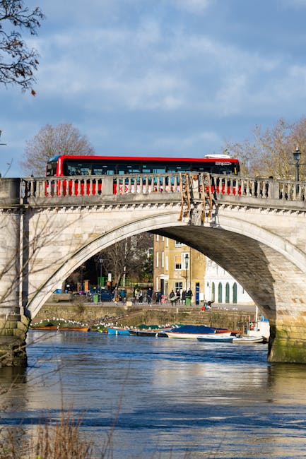 A stone arch bridge spanning over a river, with a red and black bus driving along the top. The bridge features ornate, weathered stonework with a balustrade railing painted in red and white. Beneath the bridge, the river reflects the blue sky, with small boats moored along the bank on the far side. In the background, there are trees with sparse foliage indicating a season like autumn, and a row of buildings with light-colored facades and multiple windows. The scene is illuminated by natural daylight, creating clear shadows and highlighting the textures of the stone bridge and water surface. The environment suggests an urban area near Kingston upon Thames, where private or alternative waste removal services such as rubbish collection might operate in conjunction with regular public utilities, supported by the visible maintained surroundings.