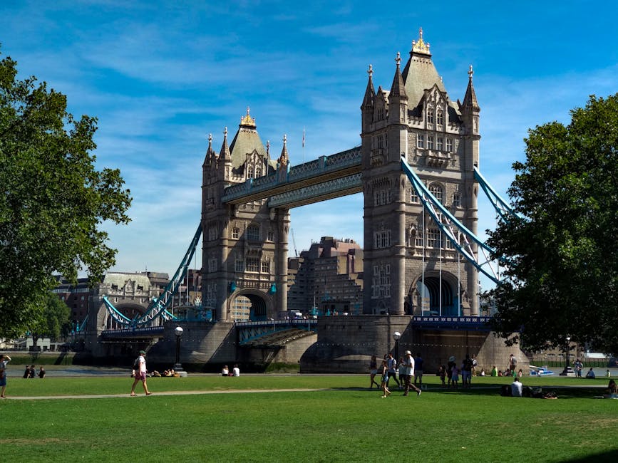 A detailed view of a historic metal bridge spanning a river in an urban environment, featuring multiple arches with ornate decorative elements and white-painted structural supports with red accents. The bridge's railing has a repeating pattern with vertical bars and ornamental details, topped with a flat walkway. In the background, there is a large, classical-style stone building with tall columns and a sloped roof, partially obscured by lush green trees. The river below shows ripples and a slightly muddy appearance, with reflections of the bridge and sky. The sky above is partly cloudy with large, white cumulus clouds and darker storm clouds, creating a contrast between bright daylight and shaded areas. The scene captures the bridge as part of an area where a private rubbish removal service, like Waste Disposal Kingston upon Thames, might facilitate alternative waste handling or on-site clearance, set against a typical cityscape backdrop.