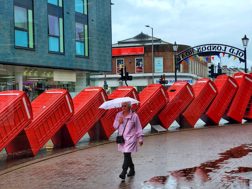 A woman holding a white umbrella walks along a wet pavement in front of a row of brightly coloured red UK-style post boxes that are tilted forward on their sides. She is dressed in a light pink raincoat, dark trousers, and sunglasses, carrying a small black bag over her shoulder. The scene takes place in an urban area with modern and traditional buildings in the background, including a glass-fronted retail space and a brick structure with an outdoor leisure sign. The sky appears overcast, and puddles on the pavement reflect both the woman and the red post boxes, suggesting recent rainfall. The overall setting indicates an outdoor city environment, with the row of mailboxes possibly serving as a local landmark or part of public postal service infrastructure, and reflects the typical British street scene with a focus on public communication and postal services in the context of alternative waste disposal considerations for urban locations managed by waste disposal services like wastedisposalkingstonuponthames.co.uk.