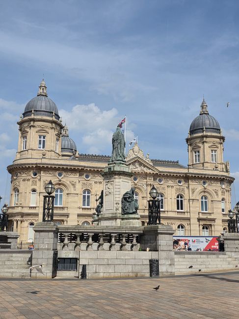 A historic stone building featuring ornate architectural details, with a central sculpture of a standing figure holding a flag atop a pedestal surrounded by smaller statues. The building has a symmetrical facade with rounded towers topped by domed roofs, decorative stonework, and evenly spaced windows. In the foreground, a paved area includes a low stone wall with black metal lamp posts and some pigeons walking across the pavement. The overall scene is captured in daylight with a partly cloudy sky in the background, exemplifying a significant public or civic building potentially related to urban infrastructure or historical landmark status, relevant to local rubbish removal or independent waste collection services for historic or government sites.